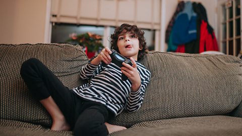 Young boy playing on a console on a sofa with his tongue sticking out