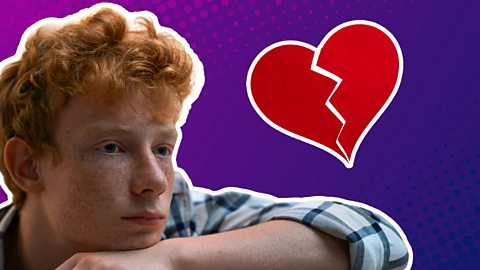 A teenage boy with ginger hair with his head on his hand looking a little down next to a broken love heart.