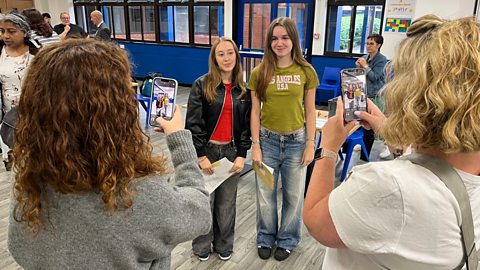 Two girls with their GCSE results stand in the foreground with their mothers taking photos of them together on their phone.