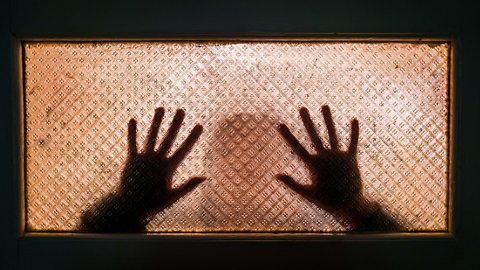 Back lit image of the silhouette of a woman with her hands pressed against a glass window. - as thought trying to escape.