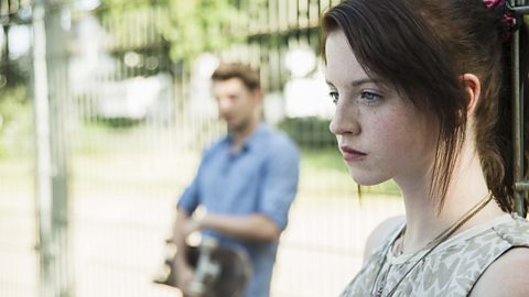 Young couple keeping their distance in a apark after an argument, he is standing against a fence holding a skateboard and she's in the foreground staring straight ahead.