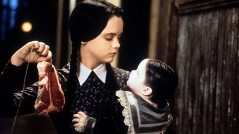 Christina Ricci plays Wednesday in film 'Addams Family Values', 1993. Wearing long black plaits, white collared dress, carrying a boy doll and a piece of raw steak