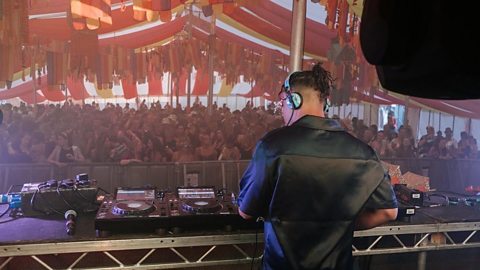 DJ wearing headphones performing on stage with turntables, mixers, and a large crowd at a music festival with vibrant banners and lights