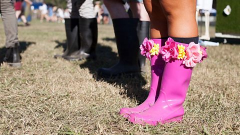 Summer festival outfits with bright pink rubber boots with colourful artificial flowers worn at an outdoor festival, surrounded by others in darker boots on dry grass