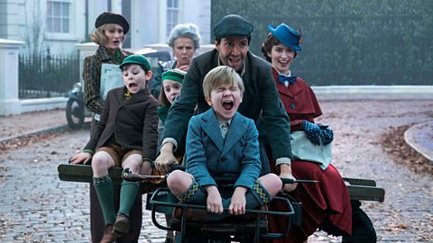 The Banks family and Mary Poppins excitedly ride along a cobbled street on a small wheeled vehcile