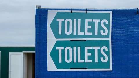 Directional signs for toilets at a music festival with hand sanitizer station and portable toilet in the background