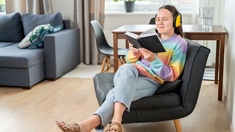 An image of a young woman relaxing in a chair with a book while wearing wireless headphones
