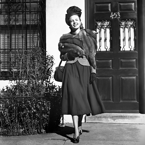 A black and white image from the 1960s of a smiling, well dressed woman leaving a house