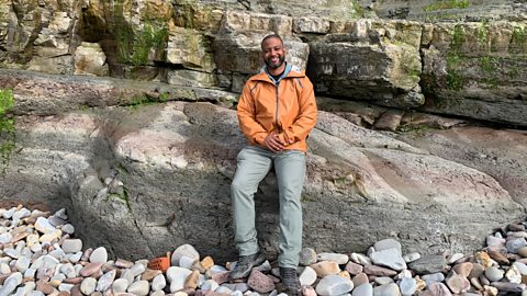 JB Gill sat on a large rock next to a rocky wall on a beach front.