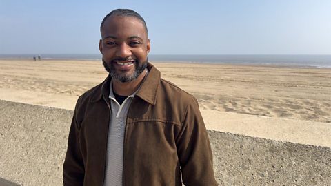 JB Gill stood next to a wall on the beach, with the shoreline against a clear blue sky in the background. 