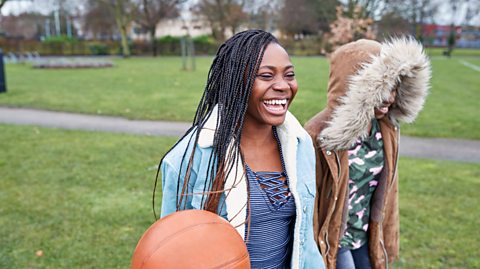 Two girls walking in coats in their local park. The one closest is laughing and holding a basketball. The girl next to her has her hood up which has thick fur on it, obscuring her face slightly.