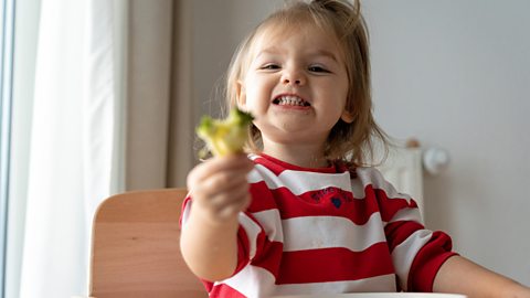 A girl grins at the camera while holding some broccoli.