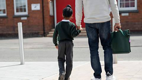 A young boy in a turban and school uniform holds his parent's hand as they approach school. The parent is holding a book bag.
