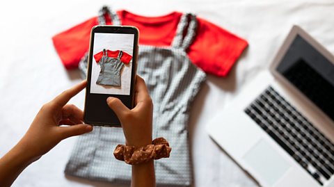 A girl uses her phone to take a photo of two items of clothing, a red tee-shirt and striped dungarees, to sell online on a nearby laptop