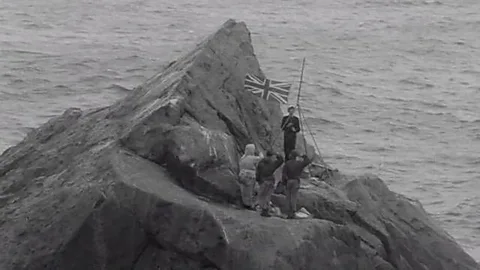 MOD Black-and-white photograph of sailors flying the British flag on Rockall (Credit: MOD)