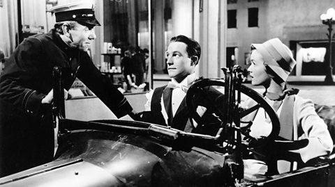 Black and white scene from the film 'Singin' in the Rain' featuring Gene Kelly as Don Lockwood driving an open-top car with Debbie Reynolds as Kathy Selden seated beside him; a uniformed officer leans into the vehicle near a storefront labeled 'Pharmacy' during a nighttime sequence.