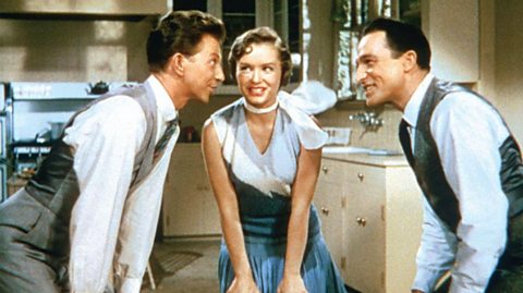 Scene from 'Singin' in the Rain' featuring Don Lockwood (Gene Kelly), Kathy Selden (Debbie Reynolds), and Cosmo Brown (Donald O'Connor) in a kitchen setting; Kathy stands center in a blue dress with white scarf, flanked by Don and Cosmo in light shirts with grey vests and ties, engaged in animated conversation.