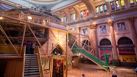 Interior of the Royal Exchange Theatre in Manchester, featuring a domed glass ceiling, ornate columns and arches; a modern exhibition structure with stairs and platforms sits on a wooden floor, with visitors exploring the historic space.