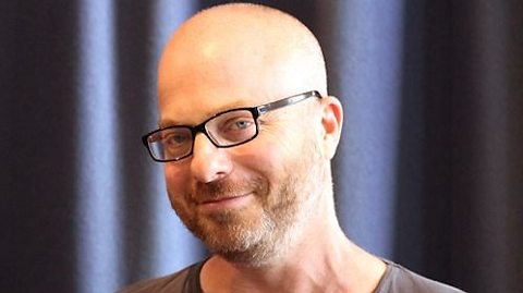 Portrait of Raz Shaw wearing a grey t-shirt, standing in front of dark curtains with a shaved head and glasses