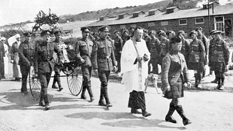  World War One soldiers in uniform walking in a funeral procession with a coffin on a wheeled cart, followed by a chaplain.