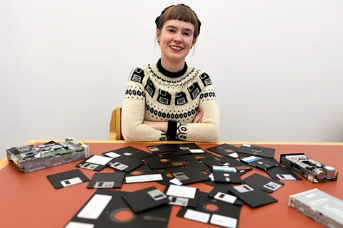 Cambridge University Library A smiling woman with her arms folded sits in front of a table with floppy disks scattered across it (Credit: Cambridge University Library)