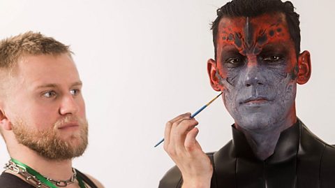 Makeup artist Axel paints a man's face red and bluey-grey to look like a monstrous creature