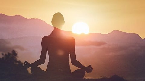 The figure of a person meditating in front of a sunrise that appears over a mountainous landscape