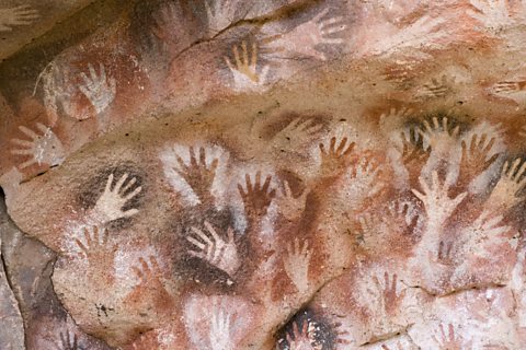 A cave wall covered in hands, where pigment has been applied around the hand to create an outline