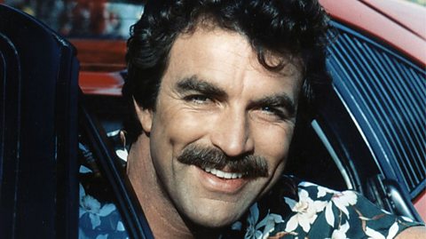 Actor Tom Selleck leans out of a red sports car wearing a hawaiian shirt, dark curly hair and a thick, bushy moustache
