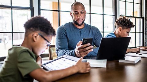 Two children sit at the table working on school work while their dad also works