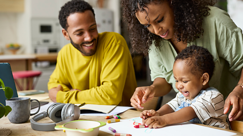 A happy toddler scribbles on paper with crayons while Mum and Dad look on.