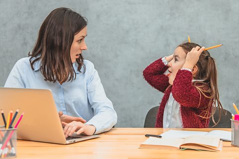 A mum and daughter at a desk, the mum tries to work, the daughter messes with pencils, smiling