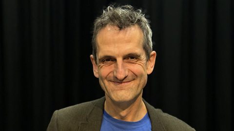 Dr Oliver Double smiling wearing a blue shirt and grey blazer