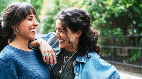 A young woman in a light denim jacket and grey top laughs while resting her right arm on the left shoulder of her friend, another young woman, who is also laughing