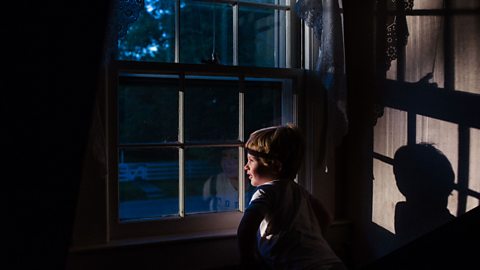 A little boy looks out of an old fashioned, single glazed window into the darkness of dusk