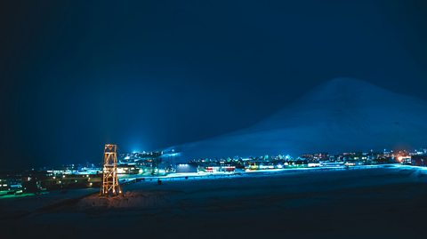 The Norwegian town of Longyearbyen in darkness. Lights are on buildings which lightly illuminate a mountain in the background.