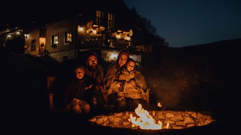 A young family are wrapped up in warm coats in front of a fire pit, with dark skies around them