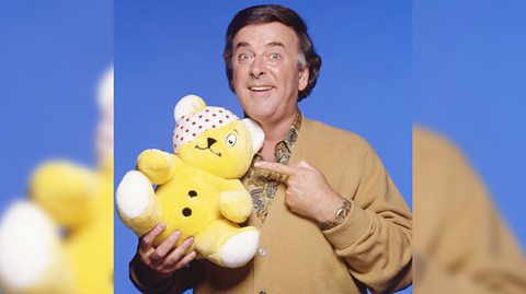 Presenter Terry Wogan smiles, holding and pointing to Pudsey Bear for Children in Need