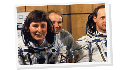 A female astronaut smiling alongside a male astronaut, with another man sitting behind them