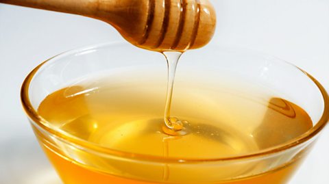 Honey drips from a from a dipper into a glass bowl.