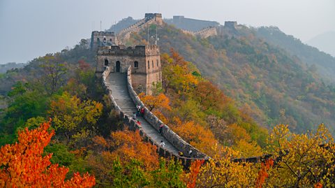 Part of the Great Wall of China seen from the distance during autumn, with orange and brown leaves on the trees surrounding it