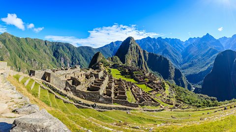 An aerial photo of Machu Picchu In the High Andes of Peru on a sunny day, showing the ruins of the lost city and the greens mountains surrounding it