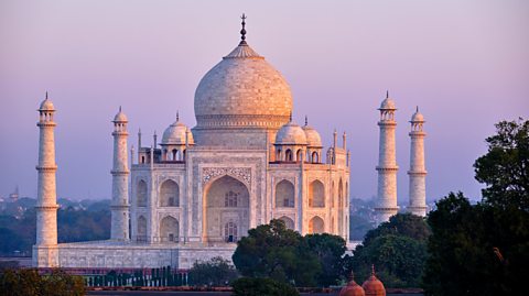 The Taj Mahal in India at sunset. The grand building is surrounded by gardens and made up of white marble, pillars and domes