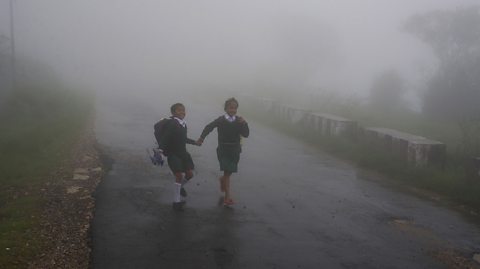 Two school children hold hands walking along a misty, rainy road