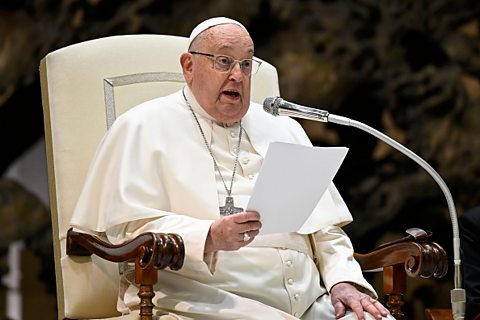 Pope Francis sitting in a large white chair reading into a microphone from a piece of paper