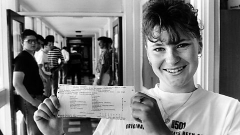 A student smiles and stands in the corridor, holding her GCSE results card in her hand