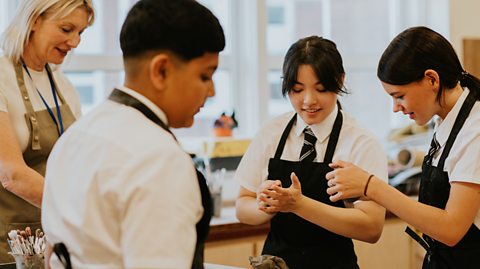A teacher and three students stand in a class room, all wearing aprons as they work on an art project with clay