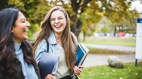 Two female students holding books and folders laugh and walk together through a campus, which is full of trees and green areas