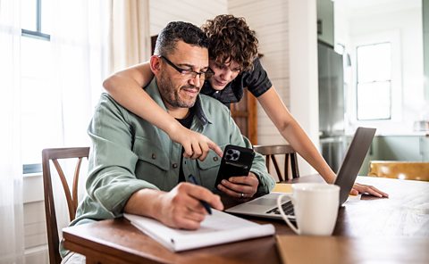 Father and son at table interacting with laptop, phone and notebook for a live lesson