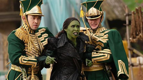 Cynthia Erivo as Elphaba in Wicked: For Good. She is being held by the Wizard's soldiers wearing green and gold uniforms and hats